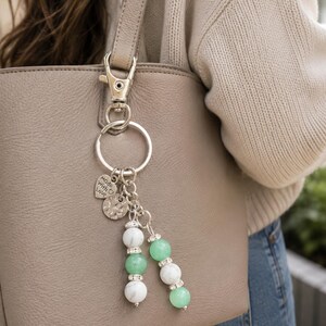 May include: A silver-toned keyring featuring a metal lobster clasp and ring, adorned with a heart-shaped charm engraved with the text made with love, a textured circular disc, and two dangling strands of white marble and green beads with rhinestones.