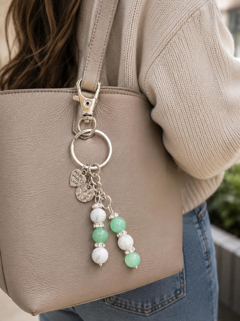 May include: A silver-toned keyring featuring a metal lobster clasp and ring, adorned with a heart-shaped charm engraved with the text made with love, a textured circular disc, and two dangling strands of white marble and green beads with rhinestones.