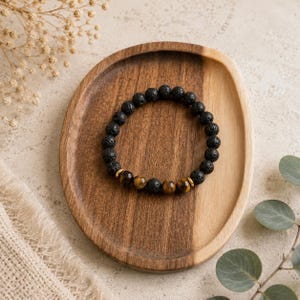 May include: A beaded bracelet featuring round, porous black lava stone beads and three smooth, golden-brown tiger eye beads. The bracelet is arranged in a circular shape, showcasing a contrast between the dark, textured volcanic rock and the polished, striped gemstone beads.