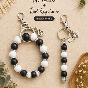 K&ouml;nnte beinhalten: Ein Set aus einem Armband-Schl&uuml;sselanh&auml;nger und einem Stab-Schl&uuml;sselanh&auml;nger mit abwechselnd mattschwarzen und wei&szlig;en Perlen sowie silberfarbenen Akzenten. Das Armband ist kreisf&ouml;rmig, der Stab-Anh&auml;nger linear. Beide haben einen Karabiner.