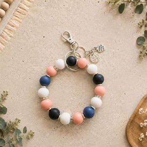 Peut inclure: Un porte-cl&eacute;s circulaire compos&eacute; de perles en silicone mat alternant bleu marine, blanc et p&ecirc;che, rehauss&eacute;es d'entretoises argent&eacute;es. Il comprend un mousqueton en m&eacute;tal argent&eacute;, un anneau bris&eacute; et deux petits pendentifs, dont un grav&eacute; Made with Love.