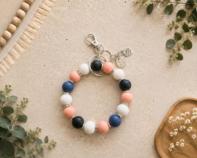 Peut inclure: Un porte-cl&eacute;s circulaire compos&eacute; de perles en silicone mat alternant bleu marine, blanc et p&ecirc;che, rehauss&eacute;es d'entretoises argent&eacute;es. Il comprend un mousqueton en m&eacute;tal argent&eacute;, un anneau bris&eacute; et deux petits pendentifs, dont un grav&eacute; Made with Love.