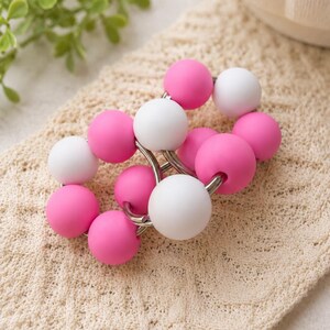 May include: A circular accessory featuring a series of alternating matte pink and white spherical beads strung together on a silver-toned metal wire frame. The beads are smooth and uniform in size, creating a continuous, interconnected loop design.