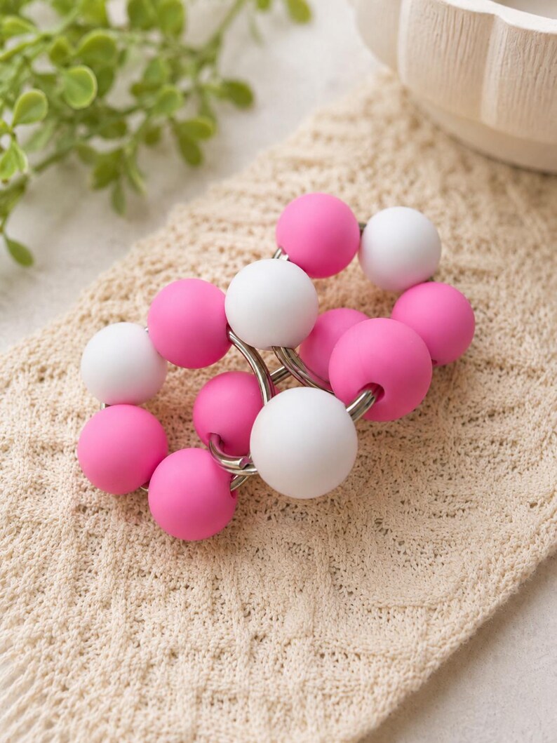 May include: A circular accessory featuring a series of alternating matte pink and white spherical beads strung together on a silver-toned metal wire frame. The beads are smooth and uniform in size, creating a continuous, interconnected loop design.