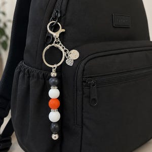 K&ouml;nnte beinhalten: Ein schwarzer Rucksack mit einem Perlen-Schl&uuml;sselanh&auml;nger am Rei&szlig;verschluss. Der Anh&auml;nger besteht aus dunkelgrauen, wei&szlig;en und orangefarbenen Perlen, die durch silberne Zwischenelemente getrennt sind, sowie einem herzf&ouml;rmigen Anh&auml;nger mit Made with Love.