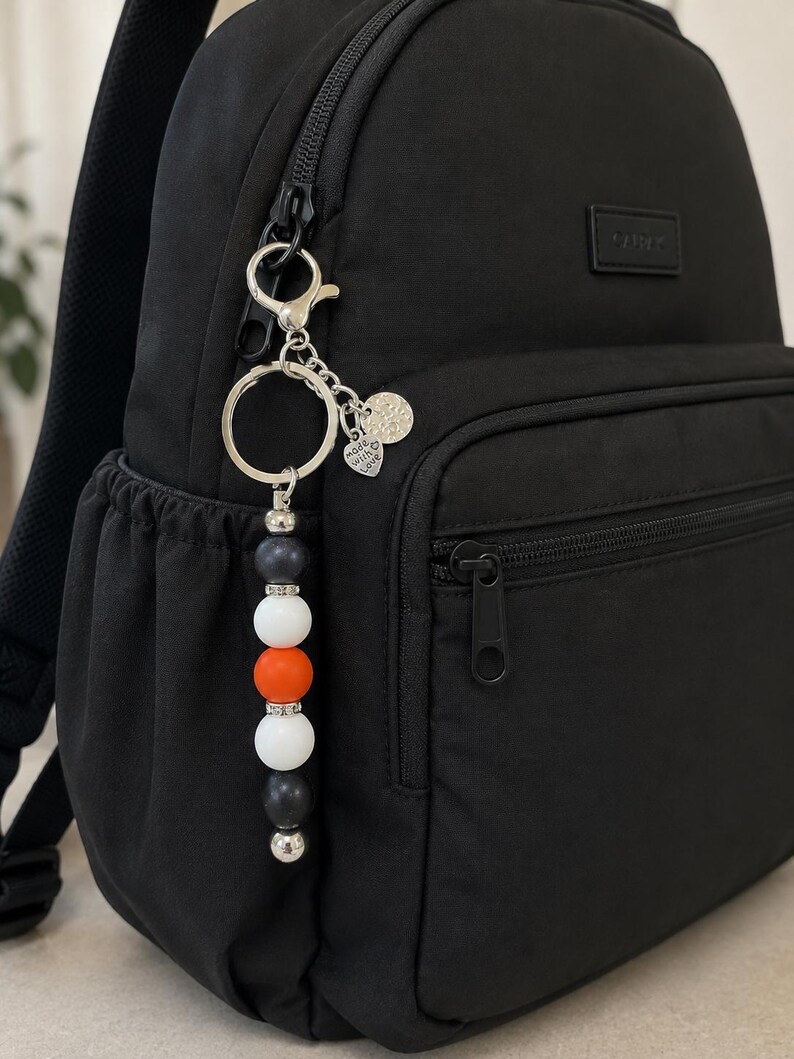 K&ouml;nnte beinhalten: Ein schwarzer Rucksack mit einem Perlen-Schl&uuml;sselanh&auml;nger am Rei&szlig;verschluss. Der Anh&auml;nger besteht aus dunkelgrauen, wei&szlig;en und orangefarbenen Perlen, die durch silberne Zwischenelemente getrennt sind, sowie einem herzf&ouml;rmigen Anh&auml;nger mit Made with Love.