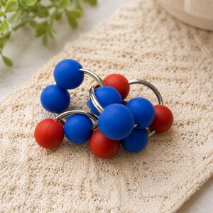 May include: A sensory toy consisting of multiple smooth, matte-finish spheres in vibrant blue and red colours. The spheres are securely attached to several interlocking silver-toned metal rings, creating a flexible, circular cluster of beads.