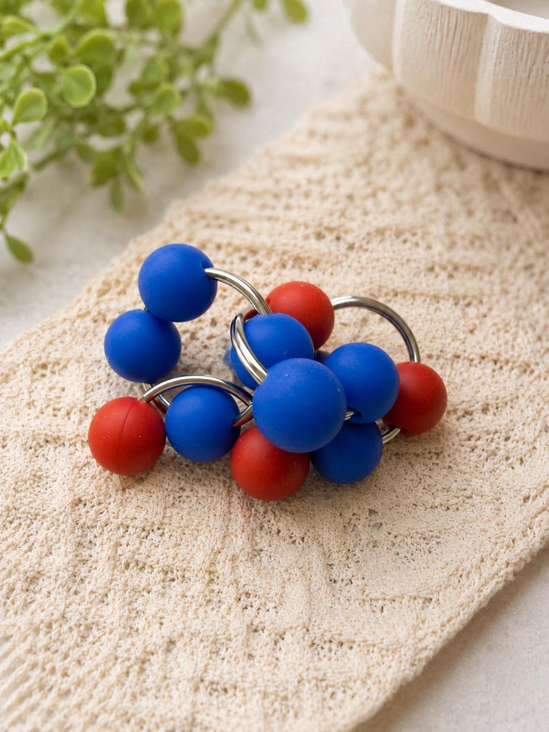 May include: A sensory toy consisting of multiple smooth, matte-finish spheres in vibrant blue and red colours. The spheres are securely attached to several interlocking silver-toned metal rings, creating a flexible, circular cluster of beads.