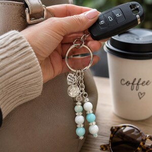 May include: A keyring featuring a silver-toned metal ring, a heart-shaped charm engraved with the text "made with love", a textured circular charm, and two dangling strands of light blue and white beads accented with small silver-toned spacers.