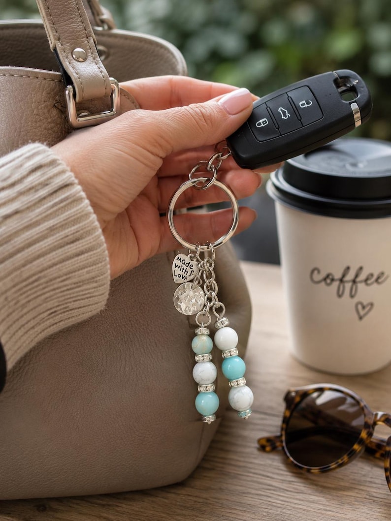 May include: A keyring featuring a silver-toned metal ring, a heart-shaped charm engraved with the text "made with love", a textured circular charm, and two dangling strands of light blue and white beads accented with small silver-toned spacers.