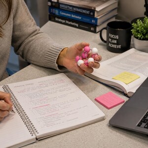 May include: A handheld fidget toy consisting of interconnected silver metal rings and alternating pink and white spherical beads. The toy is held in a palm, showcasing its circular structure and smooth, rounded components designed for tactile manipulation.