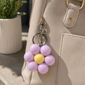 Puede incluir: Un llavero con forma de flor que presenta una esfera central amarilla rodeada por seis cuentas esf&eacute;ricas de color violeta claro. El dije est&aacute; sujeto a un mosquet&oacute;n de metal plateado y a una anilla circular, colgado de un bolso beige.