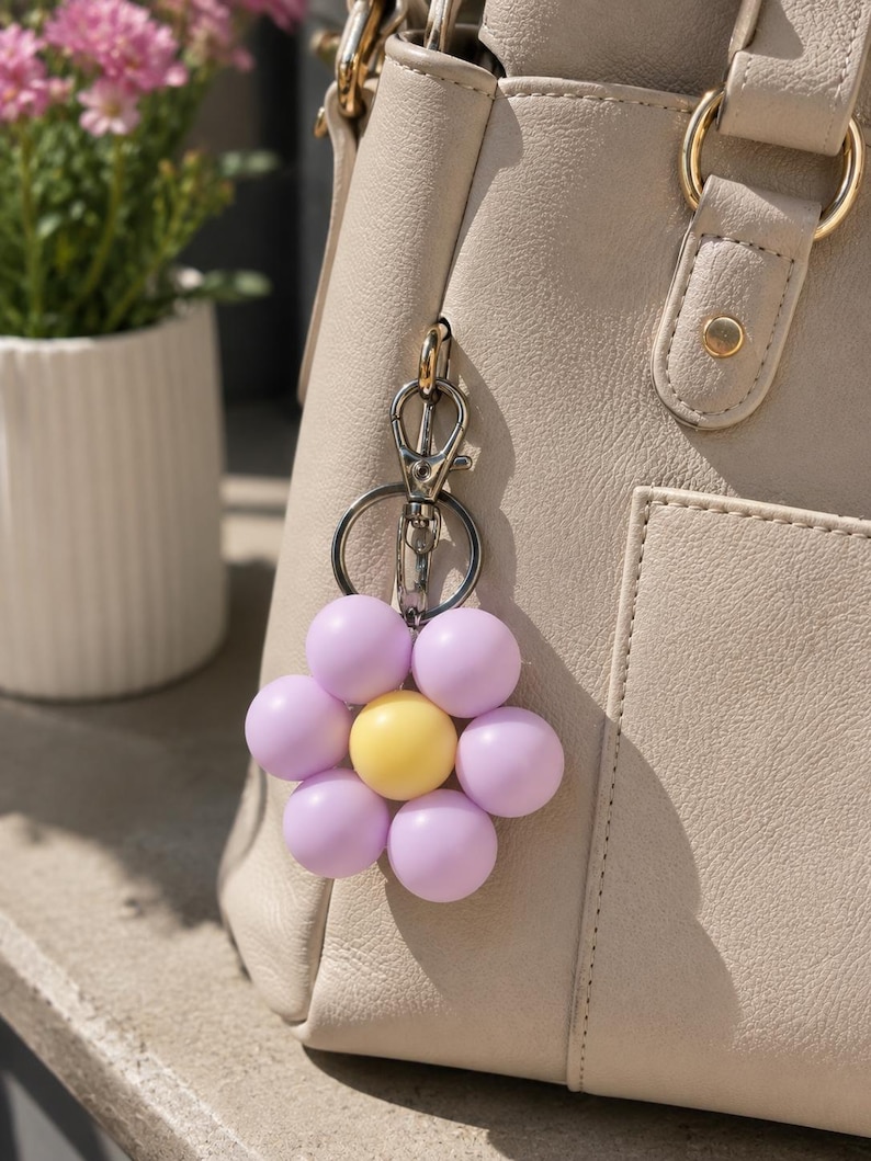 Puede incluir: Un llavero con forma de flor que presenta una esfera central amarilla rodeada por seis cuentas esf&eacute;ricas de color violeta claro. El dije est&aacute; sujeto a un mosquet&oacute;n de metal plateado y a una anilla circular, colgado de un bolso beige.