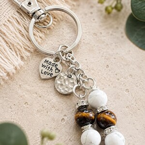 May include: A silver-toned metal keyring featuring a lobster clasp and a split ring. Attached are a heart-shaped charm with the text Made with Love, a circular hammered metal disc, and two dangling strands of white and brown tiger eye beads.