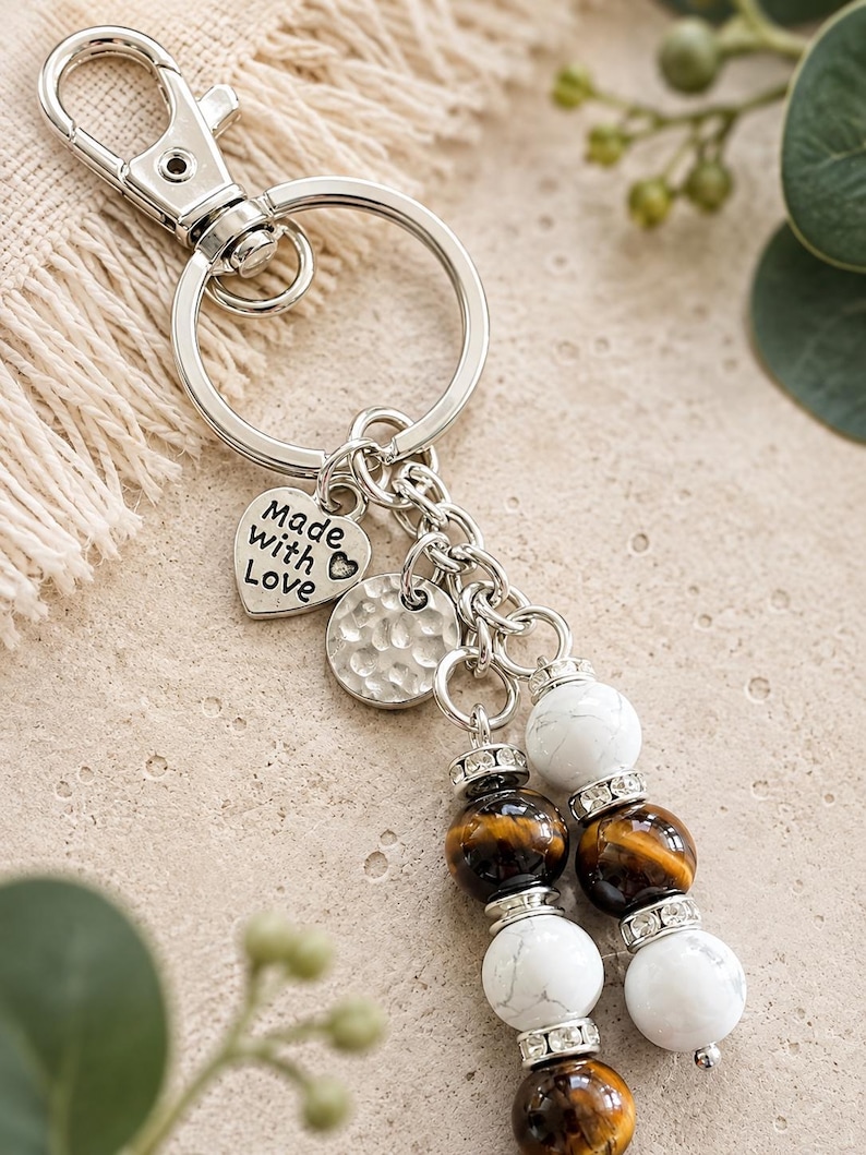 May include: A silver-toned metal keyring featuring a lobster clasp and a split ring. Attached are a heart-shaped charm with the text Made with Love, a circular hammered metal disc, and two dangling strands of white and brown tiger eye beads.