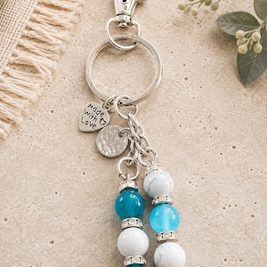 May include: A silver-toned metal keyring featuring a lobster clasp and a split ring. Attached are a heart-shaped charm engraved with the text "made with Love", a circular textured charm, and two beaded dangles with blue and white marbled beads.