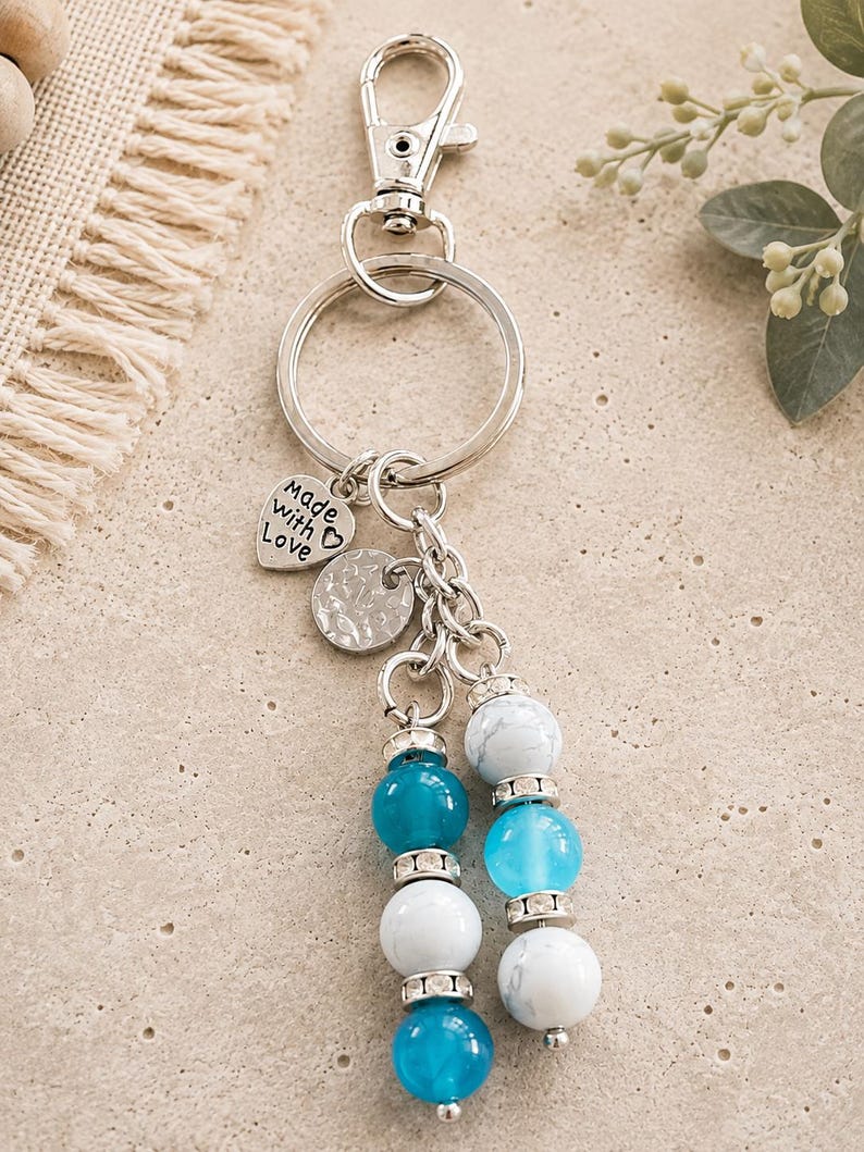 May include: A silver-toned metal keyring featuring a lobster clasp and a split ring. Attached are a heart-shaped charm engraved with the text "made with Love", a circular textured charm, and two beaded dangles with blue and white marbled beads.
