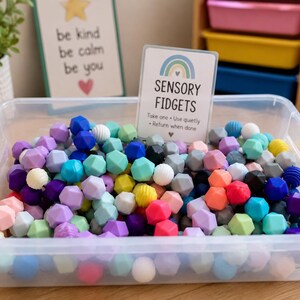 May include: A clear plastic bin filled with a variety of multi-coloured silicone geometric beads. A white sign stands in the centre with a rainbow graphic and the text SENSORY FIDGETS, Take one, Use quietly, Return when done, and a small heart.
