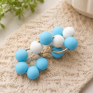 May include: A circular link accessory featuring a series of smooth, matte-finish beads in alternating shades of light blue and white. The beads are strung onto interconnected silver-toned metal rings, creating a flexible, chain-like structure.
