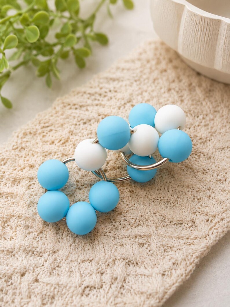 May include: A circular link accessory featuring a series of smooth, matte-finish beads in alternating shades of light blue and white. The beads are strung onto interconnected silver-toned metal rings, creating a flexible, chain-like structure.