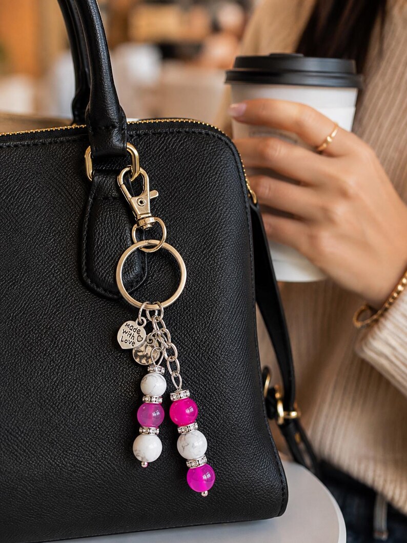 May include: A gold-toned metal keyring attached to a black textured handbag. The keyring features a round ring, a lobster clasp, and a heart-shaped charm with the text Made with Love. Two dangling strands consist of white marble-patterned beads and bright pink translucent beads separated by small silver-toned spacers with clear crystals.
