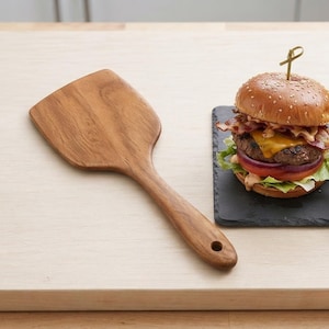 May include: A wooden spatula with a wide, flat head and a tapered handle featuring a small circular hole at the end. The kitchen utensil has a natural light brown wood grain finish and is positioned next to a cheeseburger and a side of fries.