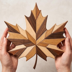 May include: A wooden maple leaf with a geometric design. The leaf is made of various shades of brown wood, including light, medium, and dark tones. The pieces are arranged in a mosaic pattern, with visible stitching along the edges. The leaf is held by two hands.