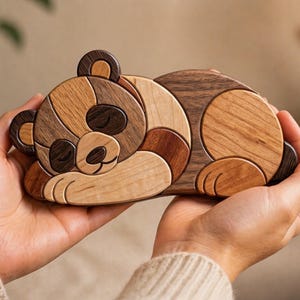 Puede incluir: Rompecabezas de madera con forma de panda durmiente. El panda está hecho de piezas de madera en diferentes tonos de marrón, beige y oscuro. Los ojos están cerrados y las patas recogidas.