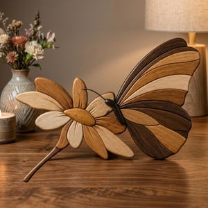Pode incluir: Uma escultura de madeira de uma borboleta e uma flor, em tons de castanho. As asas da borboleta são segmentadas com tons de madeira claros e escuros. A flor tem um centro e pétalas castanho claro. A escultura está numa superfície de madeira.