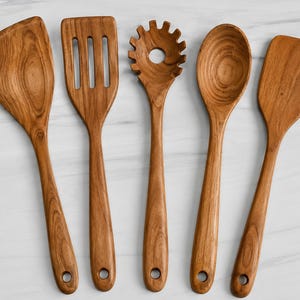 May include: A set of five wooden kitchen utensils featuring a wide angled turner, a slotted spatula, a pasta fork with a central hole, a deep round spoon, and a flat rectangular spatula, all displaying a natural brown wood grain pattern.