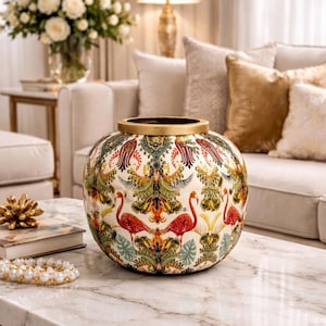 May include: A decorative ceramic vase with a gold rim and a colorful pattern featuring flamingos, birds, and foliage. The vase sits on a marble-topped table with a gold base, alongside a book and a pearl bracelet.