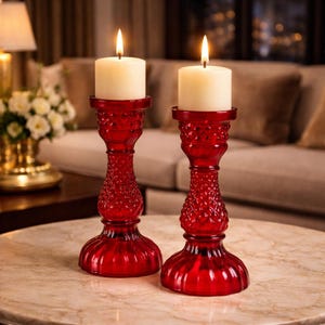 Puede incluir: Dos candelabros de vidrio rojo con velas blancas encendidas. Los candelabros tienen un patrón de diamantes texturizado en el tallo y una base redonda. Las velas son cilíndricas y brillan suavemente.