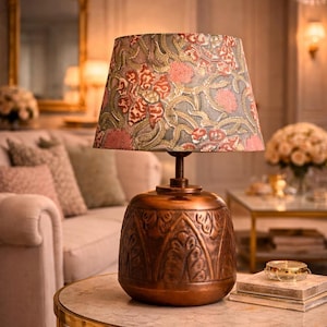 Op de afbeelding: Koperkleurige tafellamp met een bloemenpatroon lampenkap. De lampenkap heeft een ontwerp met roze en rode bloemen. De lamp staat op een tafel met een marmeren blad. De basis is gedetailleerd.