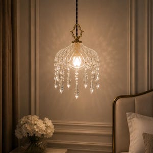 May include: A crystal chandelier with a gold-coloured frame and hanging crystal accents. The light fixture is suspended from a black cord and illuminates a bedroom setting. The chandelier has a domed shape and is decorated with multiple crystal pendants.