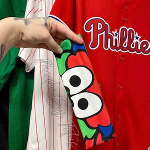 May include: Red baseball jersey with "Phillies" in white script and a star logo. The sleeve features a colorful pattern with large black and white eye shapes. Other jerseys are visible in the background.