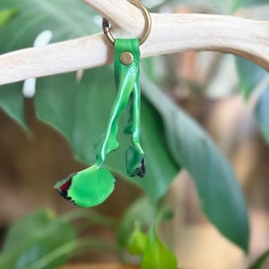 Può includere: Un portachiavi in pelle verde brillante con un design a cinturino diviso fissato con un rivetto in metallo color ottone e un anello circolare. Le estremità in pelle sono modellate come piccole foglie con bordi scuri.