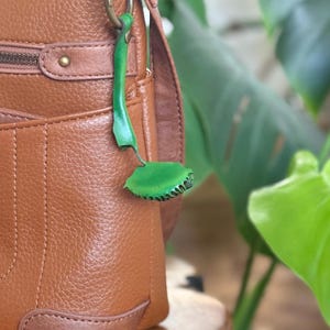 Può includere: Una borsa a tracolla in pelle martellata marrone con chiusura a zip superiore e tasca frontale, dotata di un ciondolo in pelle verde a forma di pianta attaccato all'anello in metallo color ottone sul lato dell'attacco della tracolla.