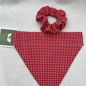 May include: A red and white gingham dog bandana and matching scrunchie. The bandana is triangular, and the scrunchie is circular. The items are made of fabric with a classic checkered pattern. A green and white tag is visible in the corner.