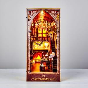 Könnte beinhalten: Ein detailliertes, beleuchtetes Miniatur-Diorama, das eine Innenszene mit Kamin, Treppe und Kronleuchter darstellt. Die Szene hat einen warmen, einladenden Glanz. Eine kleine Eulenfigur sitzt auf einem Geländer. Das Gesamtthema ist Fantasie und Miniaturkunst.