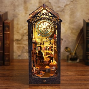 May include: A dark-toned wooden book nook insert featuring an illuminated miniature library scene. The structure includes a large clock face at the top, a desk with a chair, bookshelves, and scattered books, all lit by warm internal lighting.