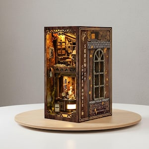 Puede incluir: Un modelo de madera iluminado de una cafetería en miniatura. El modelo presenta detalles intrincados, incluido un interior de cafetería con un barista, mesas y estanterías. El exterior tiene el texto "Serenity Coffee House" y una ventana decorativa.
