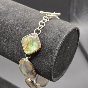 May include: A silver bracelet featuring iridescent abalone shell stones. The bracelet has a toggle clasp and a chain link design. The abalone stones display a range of colors, including green, gold, and blue. The bracelet is displayed on a black surface.