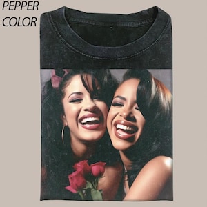 May include: A pepper-colored t-shirt featuring a graphic print of two smiling individuals with dark hair, one wearing a red hair accessory, and both positioned above a cluster of red roses. The text PEPPER COLOR is printed in the top left corner.