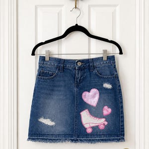 May include: A medium-wash denim skirt featuring a frayed hem, distressed patches, and embroidered appliqués. The design includes a large pink heart, a smaller pink heart with the word love, and a white roller skate with pink wheels and accents.