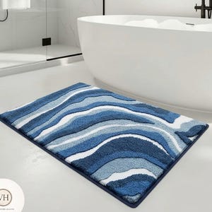 Op de afbeelding: Een rechthoekige badmat met een golvend patroon in tinten blauw en wit. De mat heeft een donkerblauwe rand en ligt op een witte vloer in een badkamer. Het ontwerp lijkt op oceaangolven.