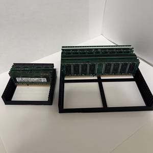 May include: Two black 3D-printed RAM holders, one small and one large, each holding multiple green RAM sticks. The holders are rectangular with a grid-like design. The background is plain white.