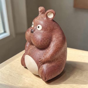 May include: A brown ceramic figurine of a cartoon-like animal with large cheeks and a white belly. The figurine has white accents on the ears and eyes, and is sitting on a wooden surface.