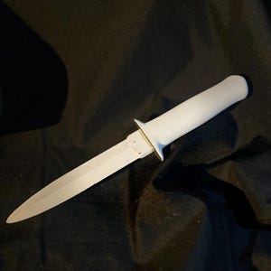 Könnte beinhalten: Ein weißer Dolch mit einer spitzen Klinge und einem weißen Griff. Die Klinge ist silberfarben und der Griff ist einfarbig weiß. Der Dolch hat einen kleinen Parier, wo die Klinge auf den Griff trifft. Der Hintergrund ist ein dunkler, strukturierter Stoff.