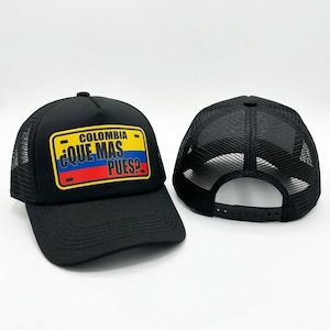 May include: Black trucker hat with a mesh back and a curved brim. The front features a rectangular patch with the word "COLOMBIA" above the phrase "¿QUE MAS PUES?" in yellow, blue, and red, resembling a license plate.