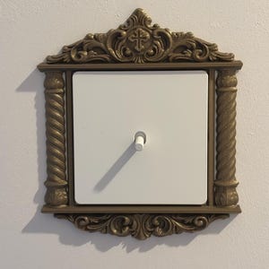 May include: An ornate, gold-coloured light switch cover with a white switch and a decorative frame. The frame features a cross at the top and scrolled details. The switch is centered on a white square.